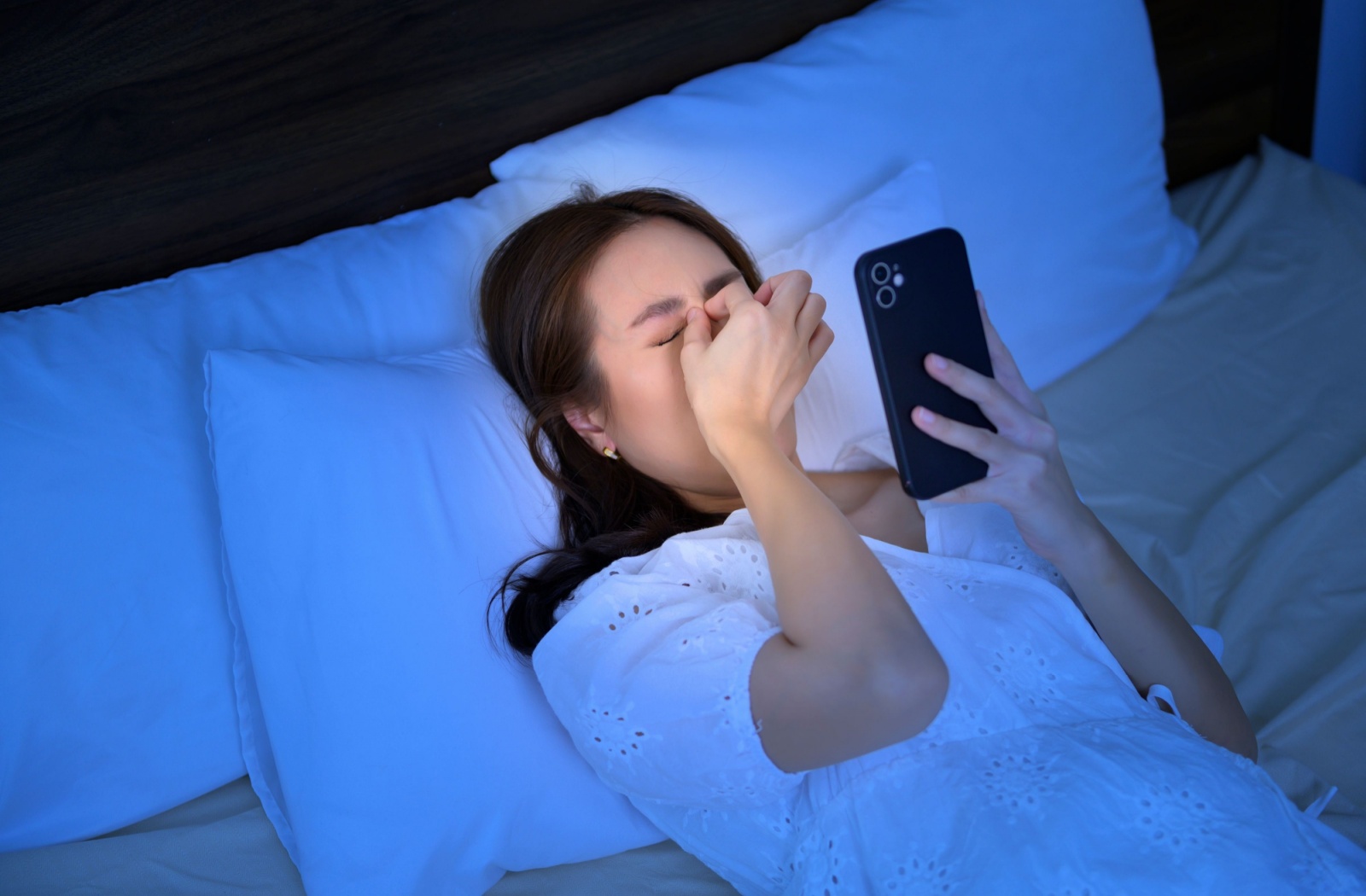 A woman places her fingers on her eyes due to discomfort from looking at her smartphone at night.