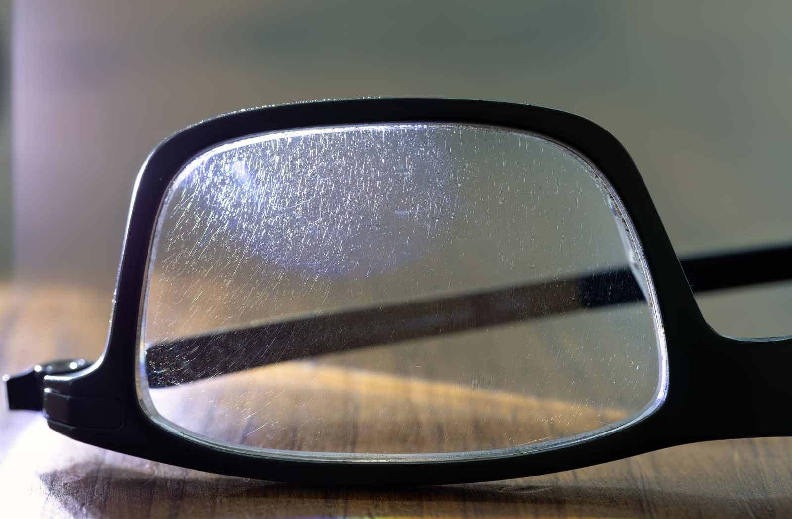 Close-up of a single lens on black eyeglasses. Sunlight illuminates many tiny scratches on the lens.