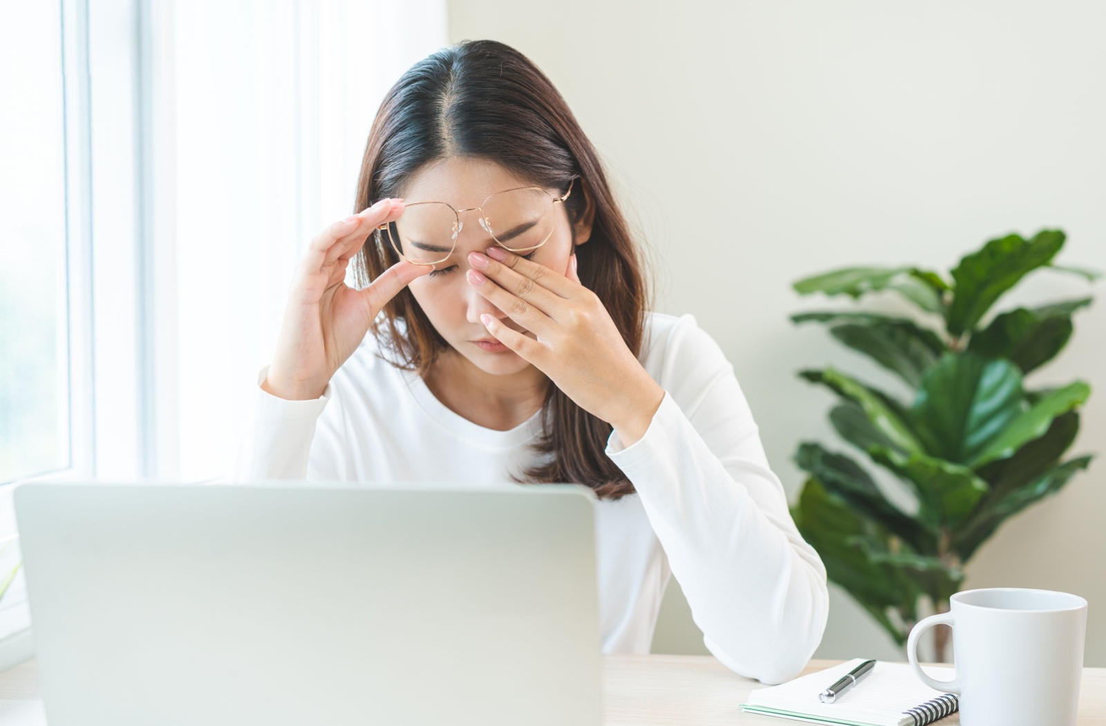 A person working on a laptop lifts their glasses and rubs their closed eye.