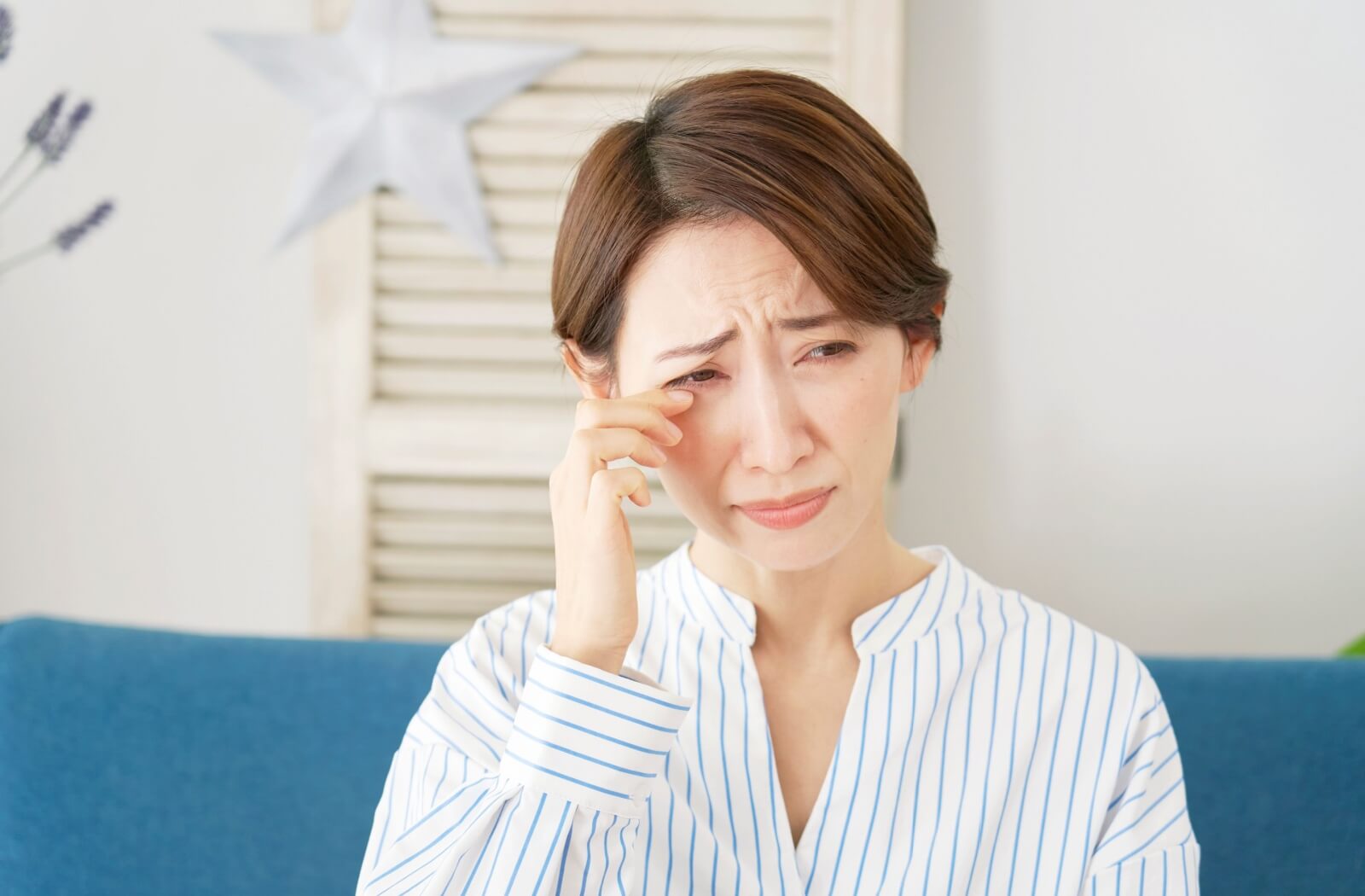 Woman sitting indoors gently rubbing her irritated eye, appearing uncomfortable, experiencing symptoms of dry eye.