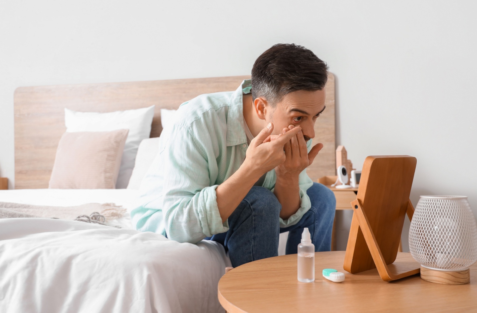 Person sitting on bed putting in a contact lens using a mirror.