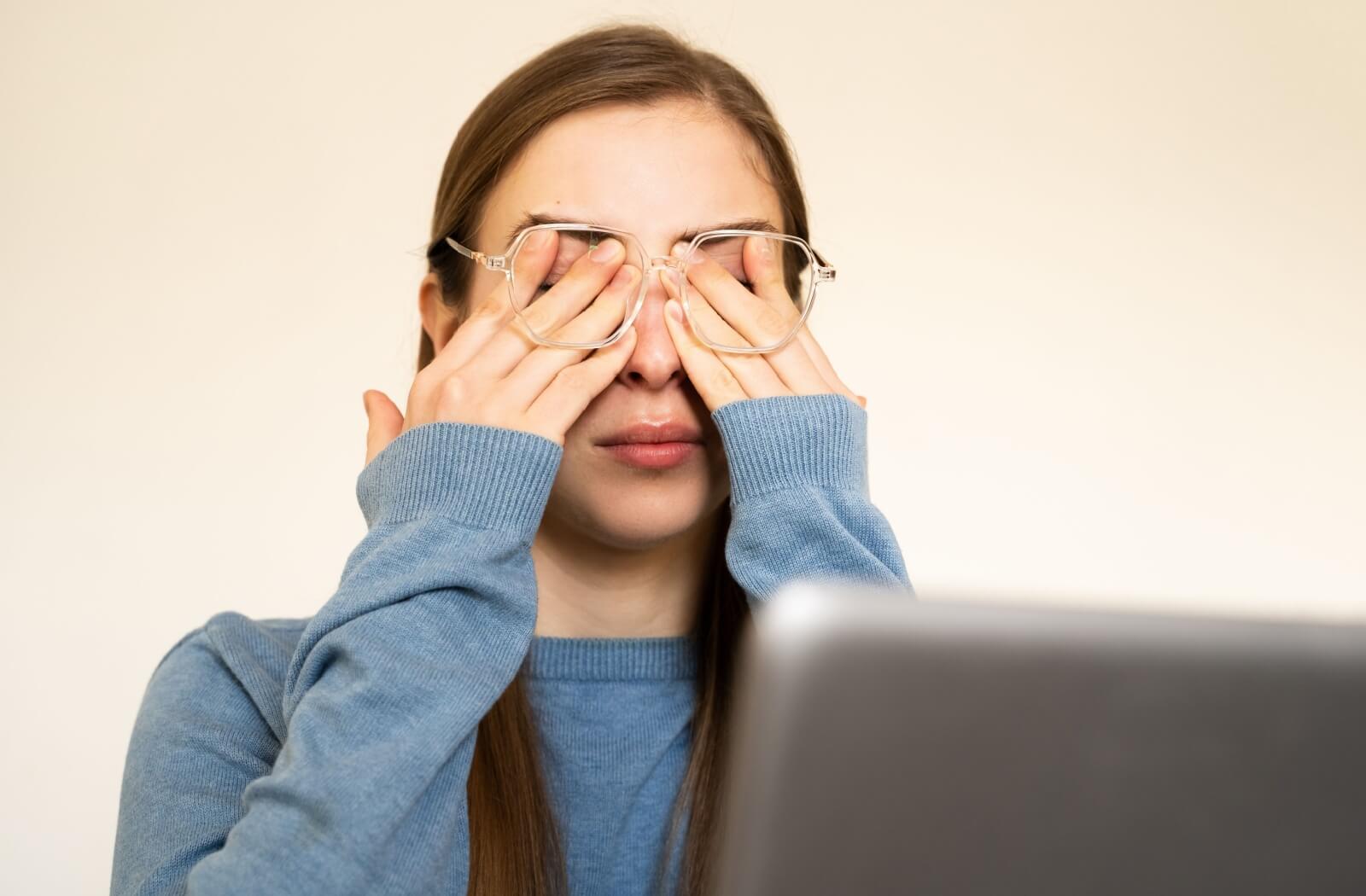 A young woman in a blue sweater rubbing her eyes underneath her clear-framed eyeglasses while sitting in front of a laptop.