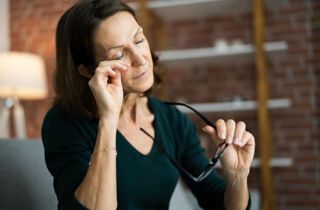 A woman holding her eyeglasses while rubbing her closed eye in discomfort indoors.